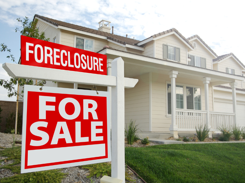 Modern beige two-story house with red "Foreclosure" and "For Sale" signs in front yard.