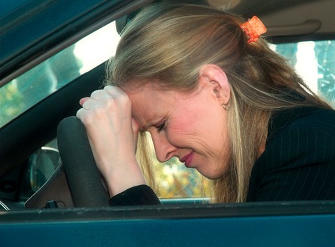Woman with blonde hair and black jacket resting her forehead on a car steering wheel.