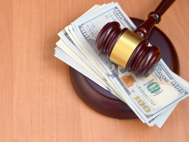 Wooden judge's gavel resting on a stack of US hundred-dollar bills on a wooden surface.