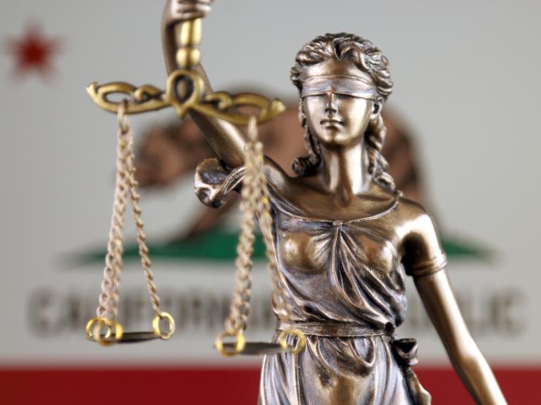 Bronze statue of Lady Justice blindfolded holding scales, with California state flag blurred in background.