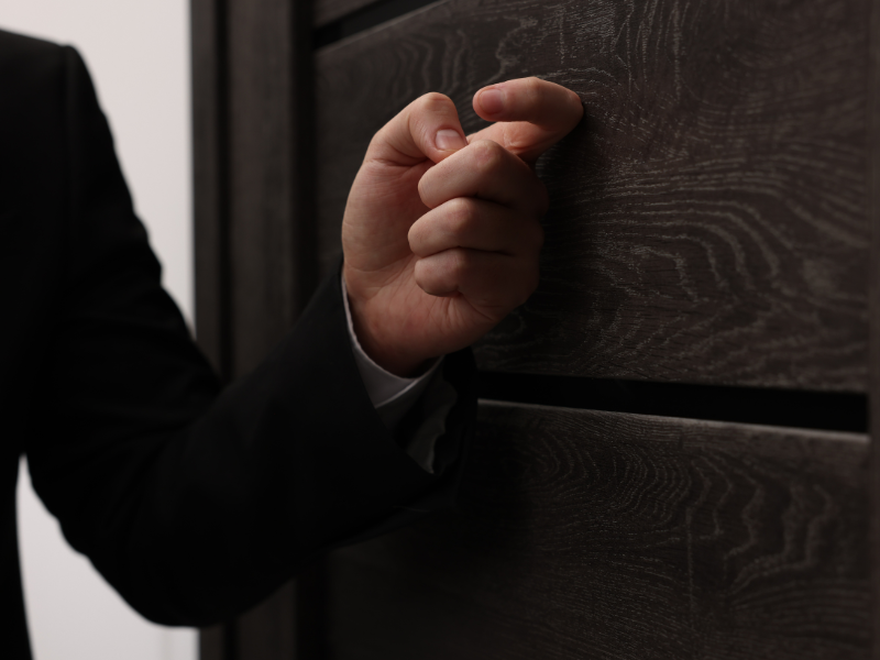 Close-up of a person in a black suit knocking on a dark wooden door.