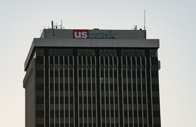 Top portion of a tall office building with a "us bank" sign in red and blue lettering against a light sky background.