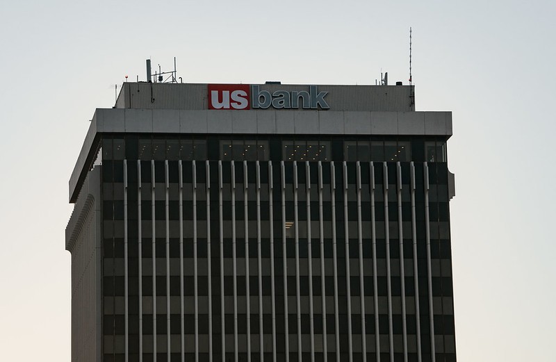 Top portion of a tall office building with a "us bank" sign in red and blue lettering against a light sky background.