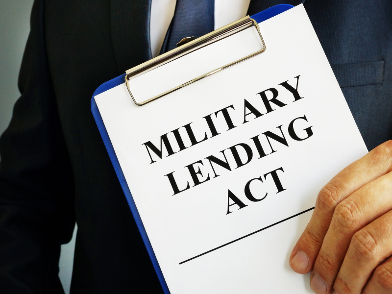 Person in dark suit holding clipboard with document titled "MILITARY LENDING ACT" in bold black letters.