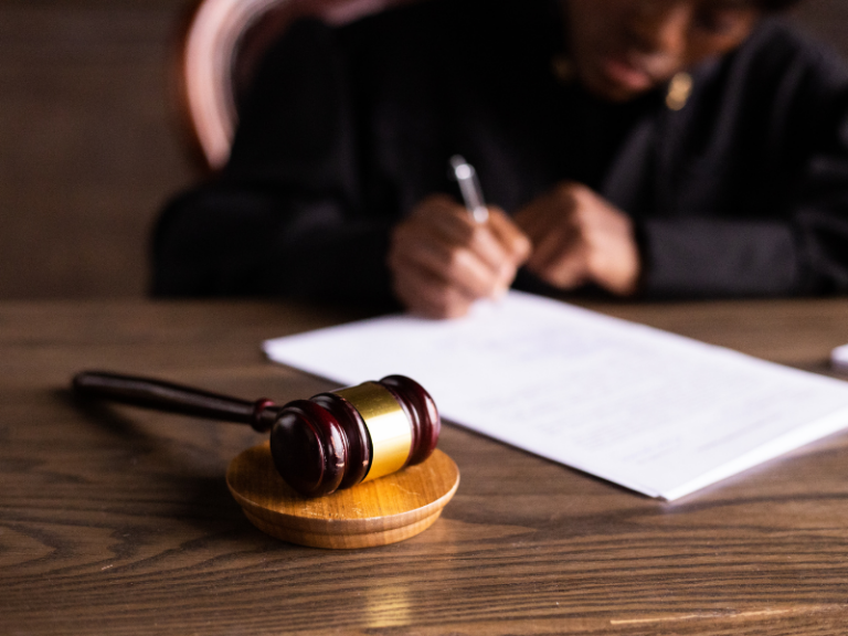 Wooden judge's gavel on desk with blurred person writing on documents in background.
