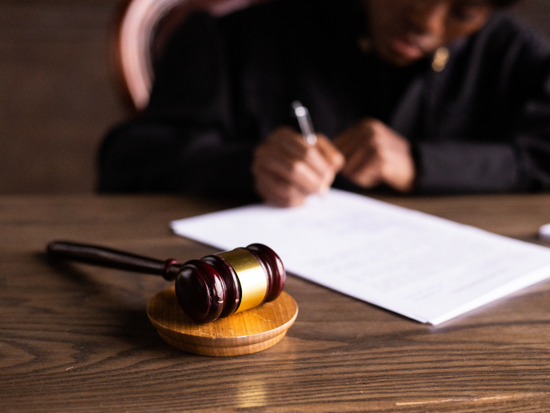 Wooden judge's gavel on desk with blurred person writing on documents in background.