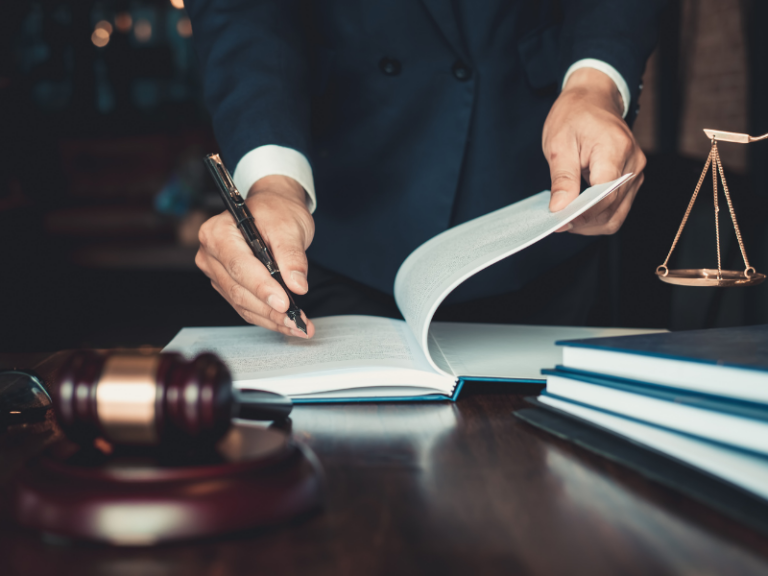 Person in dark suit reviewing legal documents at desk with gavel and scales of justice.