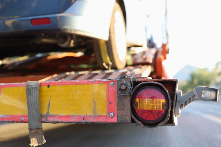 Close-up view of the rear section of a red vehicle transport trailer with a blue car loaded, highlighting the taillight and yellow reflective strip.