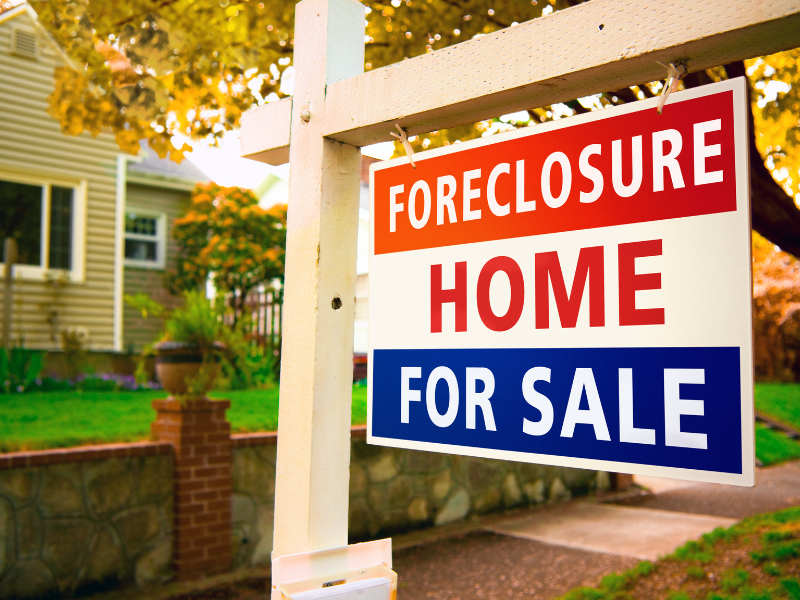 Foreclosure home for sale sign in front of suburban house with autumn foliage.