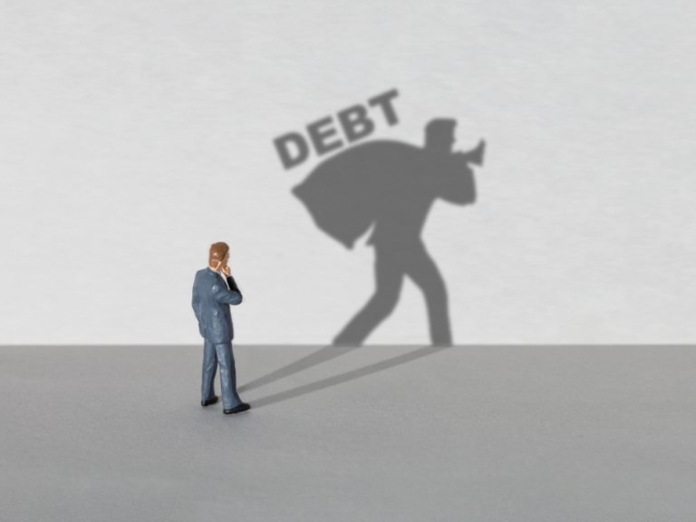 Small figurine of a man in a suit with shadow carrying a sack labeled "DEBT" on a gray surface.