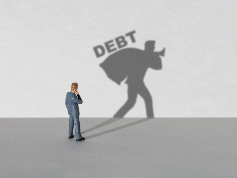 Small figurine of a man in a suit with shadow carrying a sack labeled "DEBT" on a gray surface.