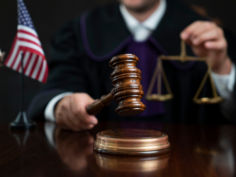 Judge in black robe holding wooden gavel and brass scales with American flag on desk.