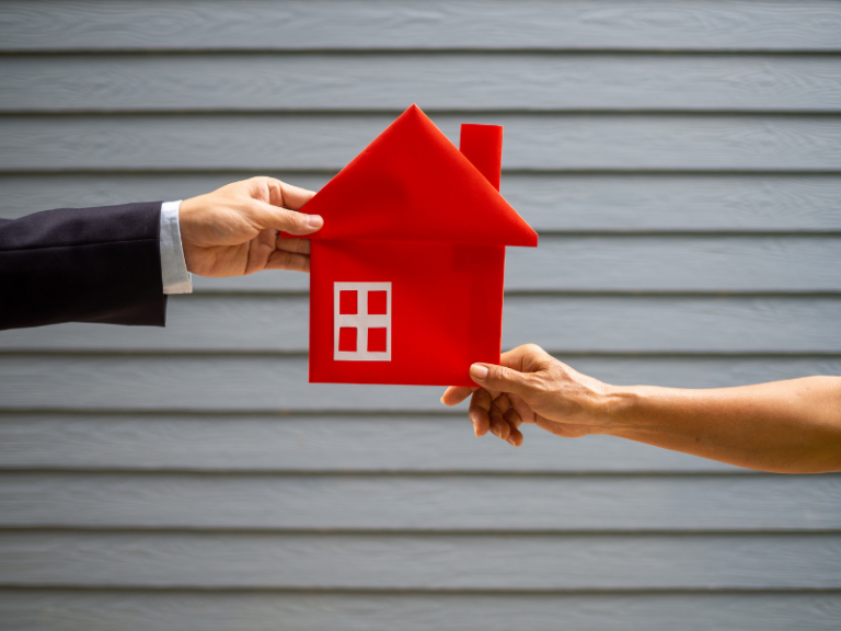 Two hands exchanging a red paper house model against a gray wooden background.