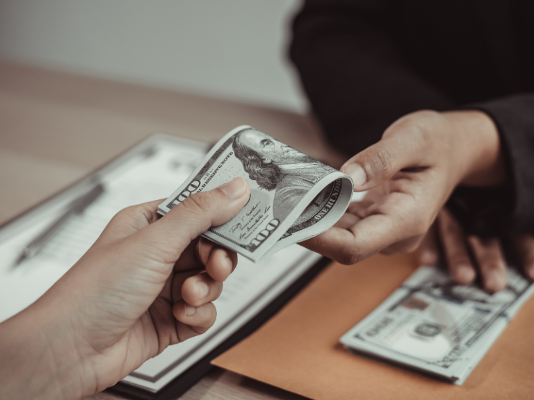 Two hands exchanging folded US hundred-dollar bills over a desk with documents and an envelope.