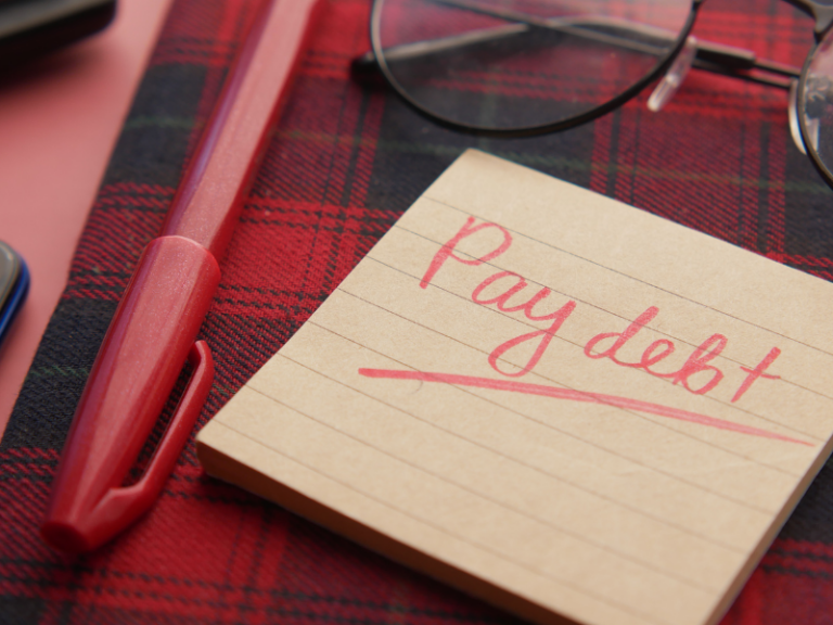 Close-up of a red pen, eyeglasses, and a note reading "Pay debt" on a red and black plaid surface.