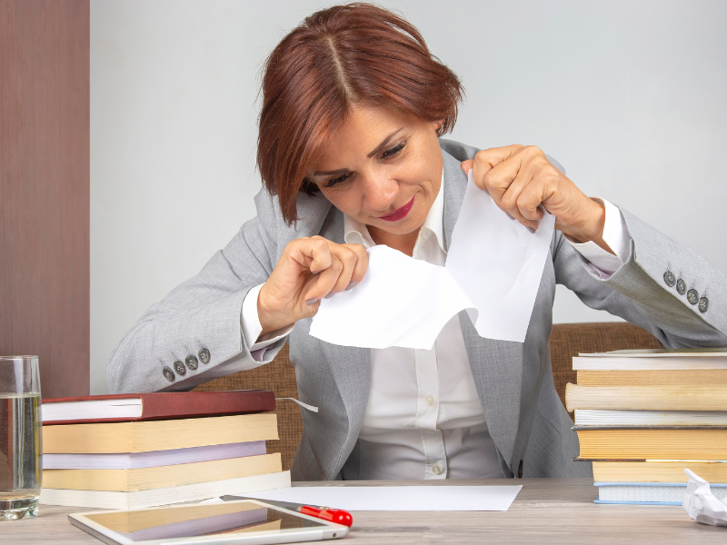 Professional woman in gray suit tearing a sheet of white paper at a desk with books and office supplies.