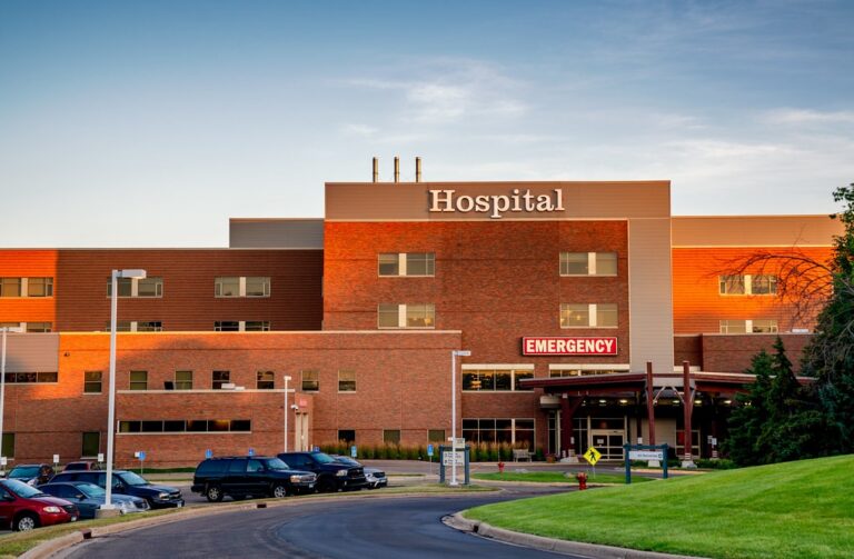 Exterior view of a brick hospital building with an emergency entrance and parked vehicles.