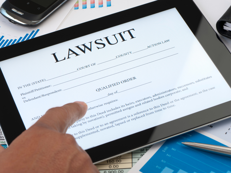 Finger pointing at a digital tablet displaying a lawsuit document, surrounded by charts and a smartphone.