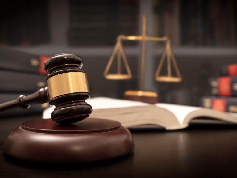 Wooden judge's gavel with brass band, open law book, and brass scales of justice.