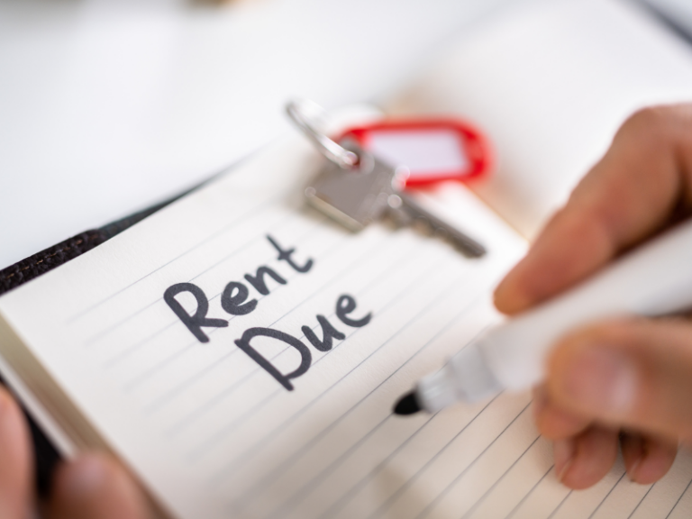 Close-up of a hand writing "Rent Due" in a notebook with house keys nearby.