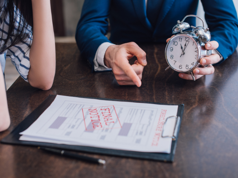 Two people at a table with a foreclosure notice and a man holding a silver alarm clock.