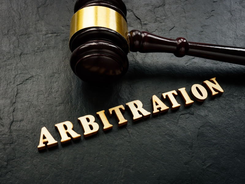 Wooden judge's gavel with gold band placed above the word "ARBITRATION" in wooden letters on dark surface.