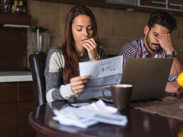 Woman reading a document at a table with a laptop and a stressed man in the background.