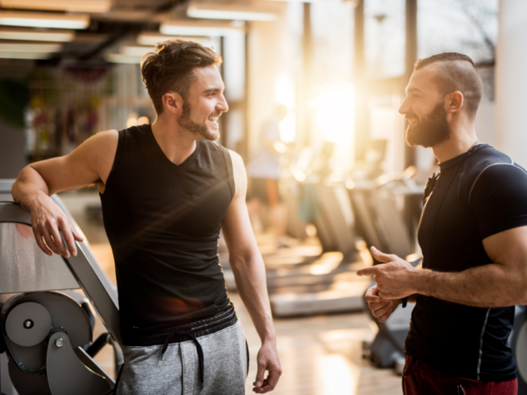 Two men in athletic wear conversing in a sunlit gym with exercise equipment in the background.