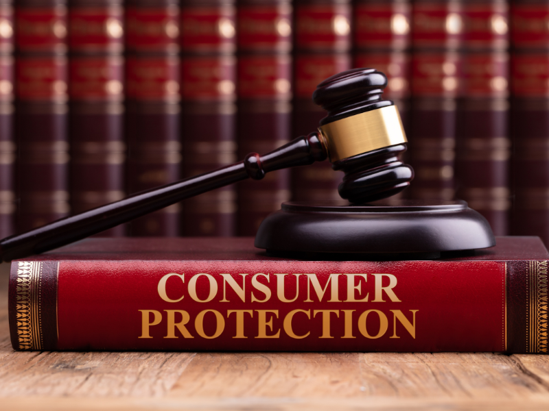 Wooden judge's gavel resting on a red book titled "Consumer Protection" with legal books in the background.