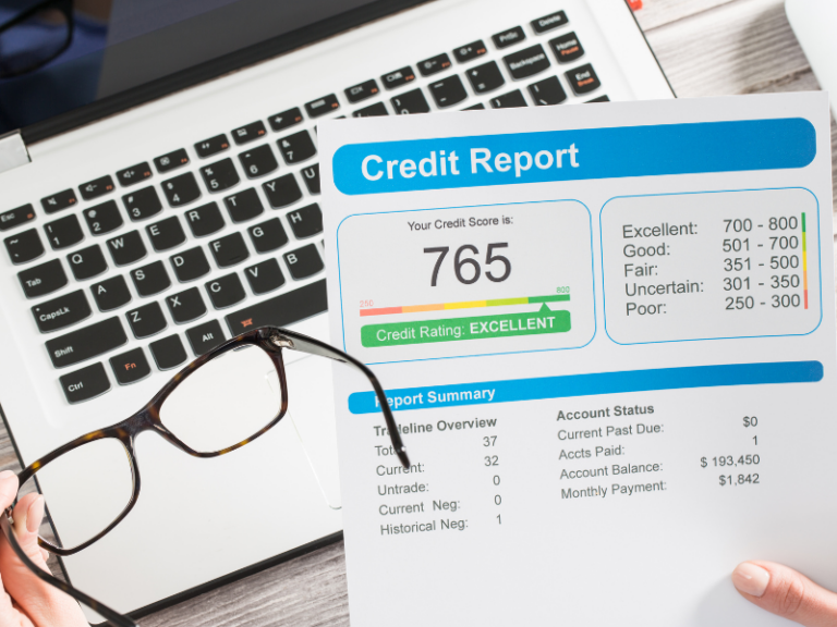 Credit report with score 765 rated excellent, held over laptop keyboard and glasses.