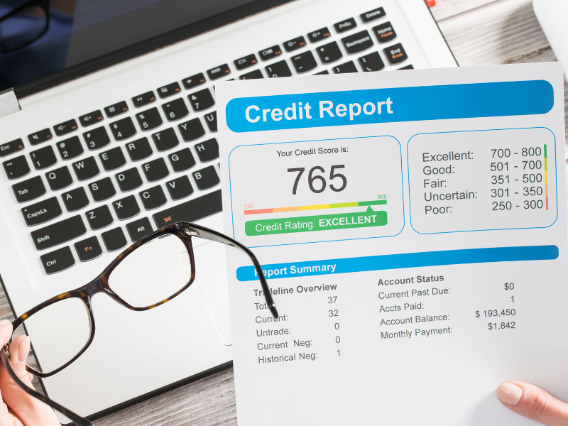 Credit report with score 765 rated excellent, held over laptop keyboard and glasses.