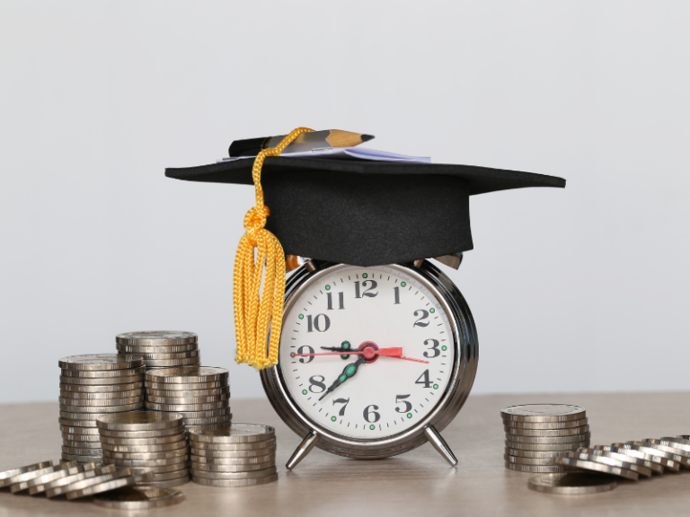 Silver alarm clock wearing a black graduation cap with a yellow tassel, surrounded by stacks of coins.