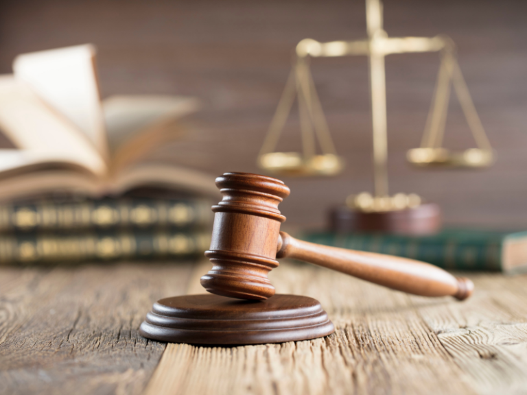 Wooden judge's gavel on a wooden table with legal books and brass scales of justice in the background.
