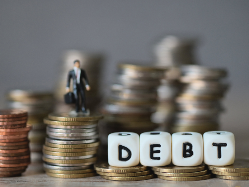 Stacked coins with a miniature businessman figurine and dice spelling "DEBT" on coins.