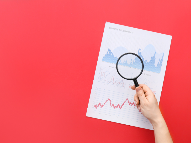Hand holding magnifying glass over business infographic charts on white paper, red background.