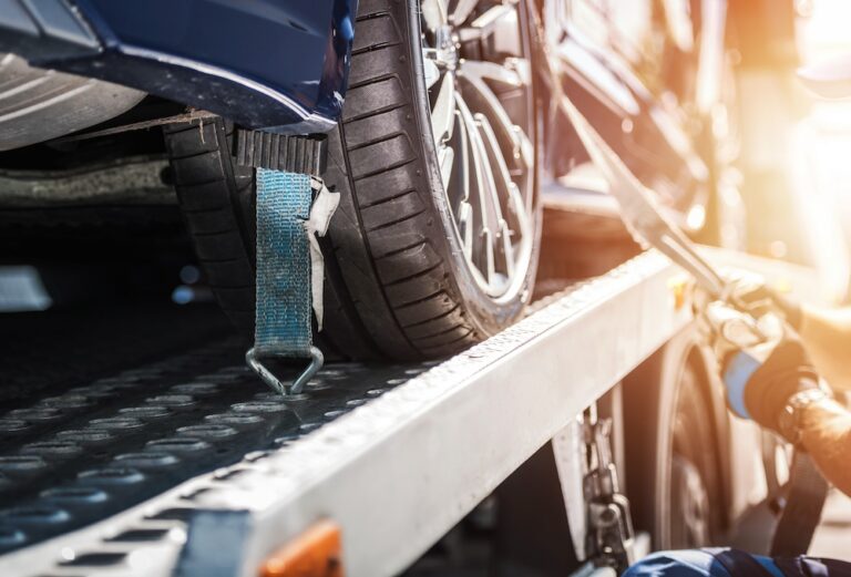 Close-up of a car tire secured with a blue strap on a metal tow truck bed, with a gloved hand tightening the strap.