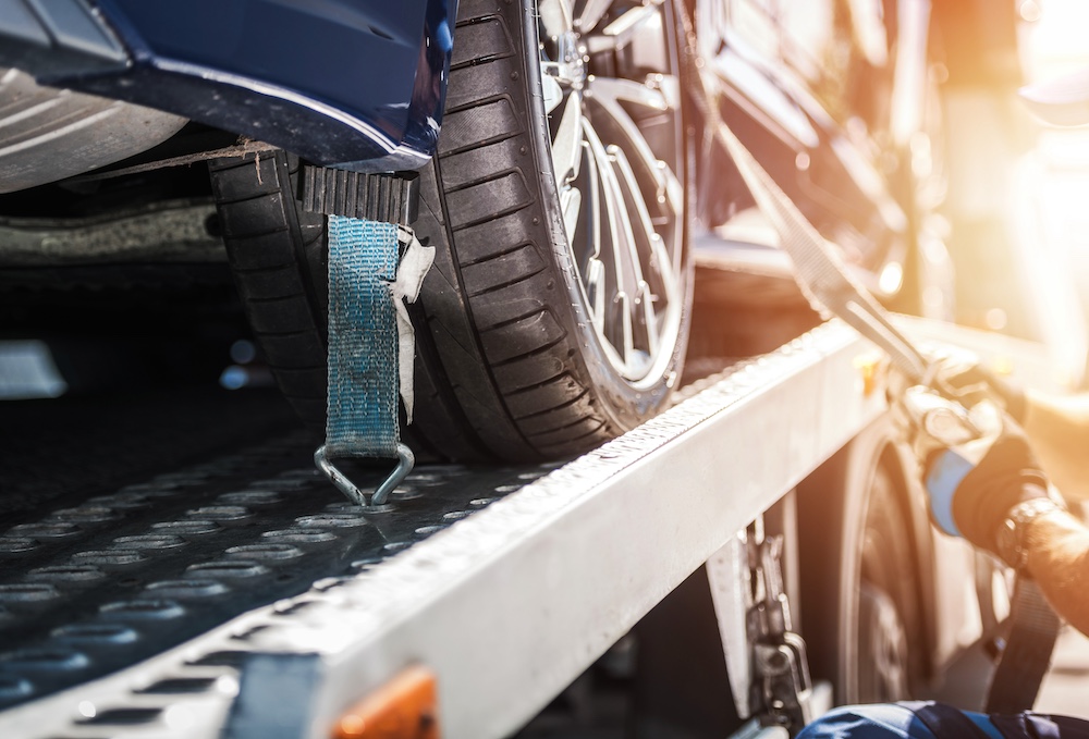 Close-up of a car tire secured with a blue strap on a metal tow truck bed, with a gloved hand tightening the strap.