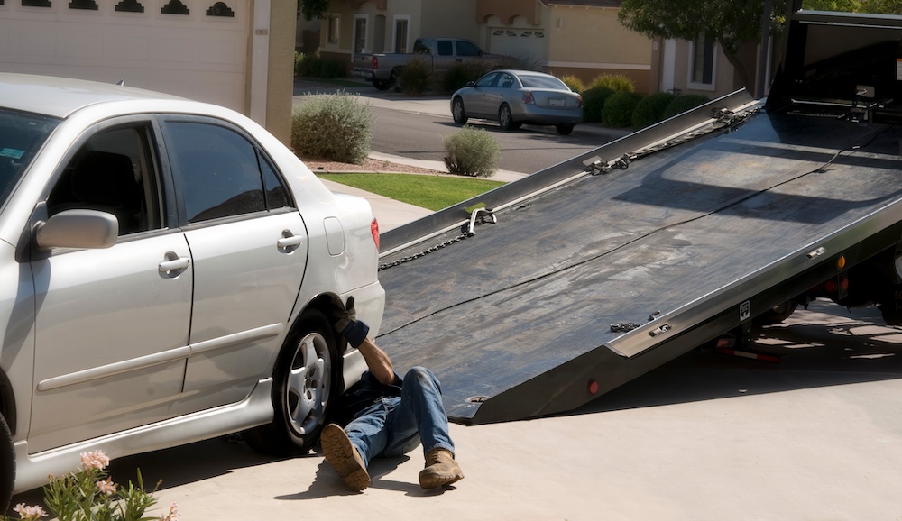 Mechanic working under a white sedan car beside a lowered flatbed tow truck in a residential driveway.