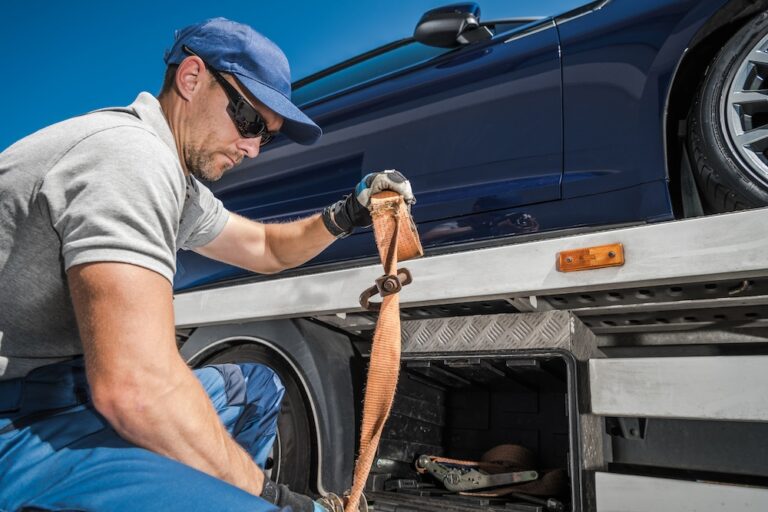 Professional male worker securing orange strap on blue car loaded on flatbed tow truck.