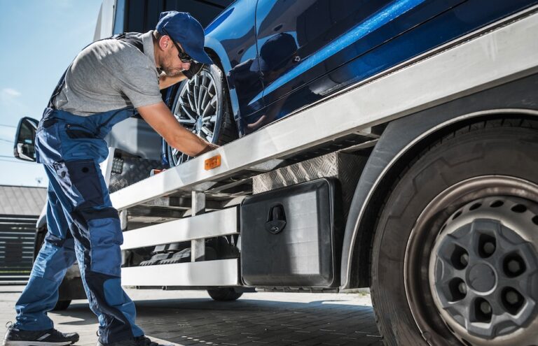 Mechanic in blue uniform securing blue car on flatbed tow truck under clear sky.