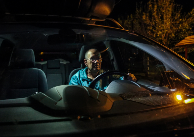 Elderly man wearing glasses driving a car at night, illuminated by dashboard light.