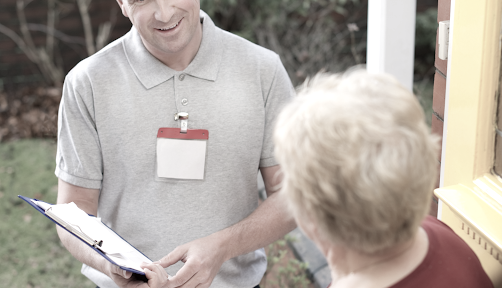 Man in gray polo shirt with badge holding clipboard, speaking to elderly woman at door.