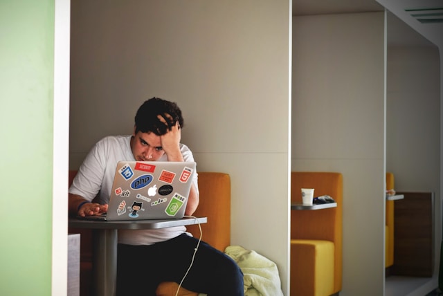 Young man in white shirt sitting at table with laptop covered in programming stickers, appearing stressed.