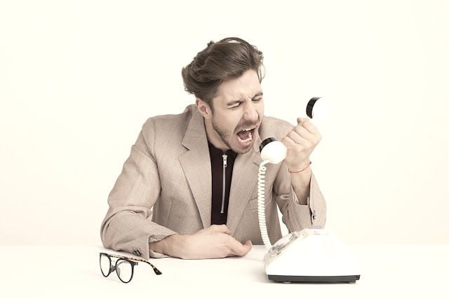 Man in beige blazer angrily shouting into a white rotary phone handset at a white desk.