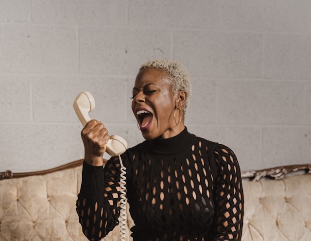 Woman with short blonde hair in a black patterned top holding a beige telephone receiver, appearing to shout.