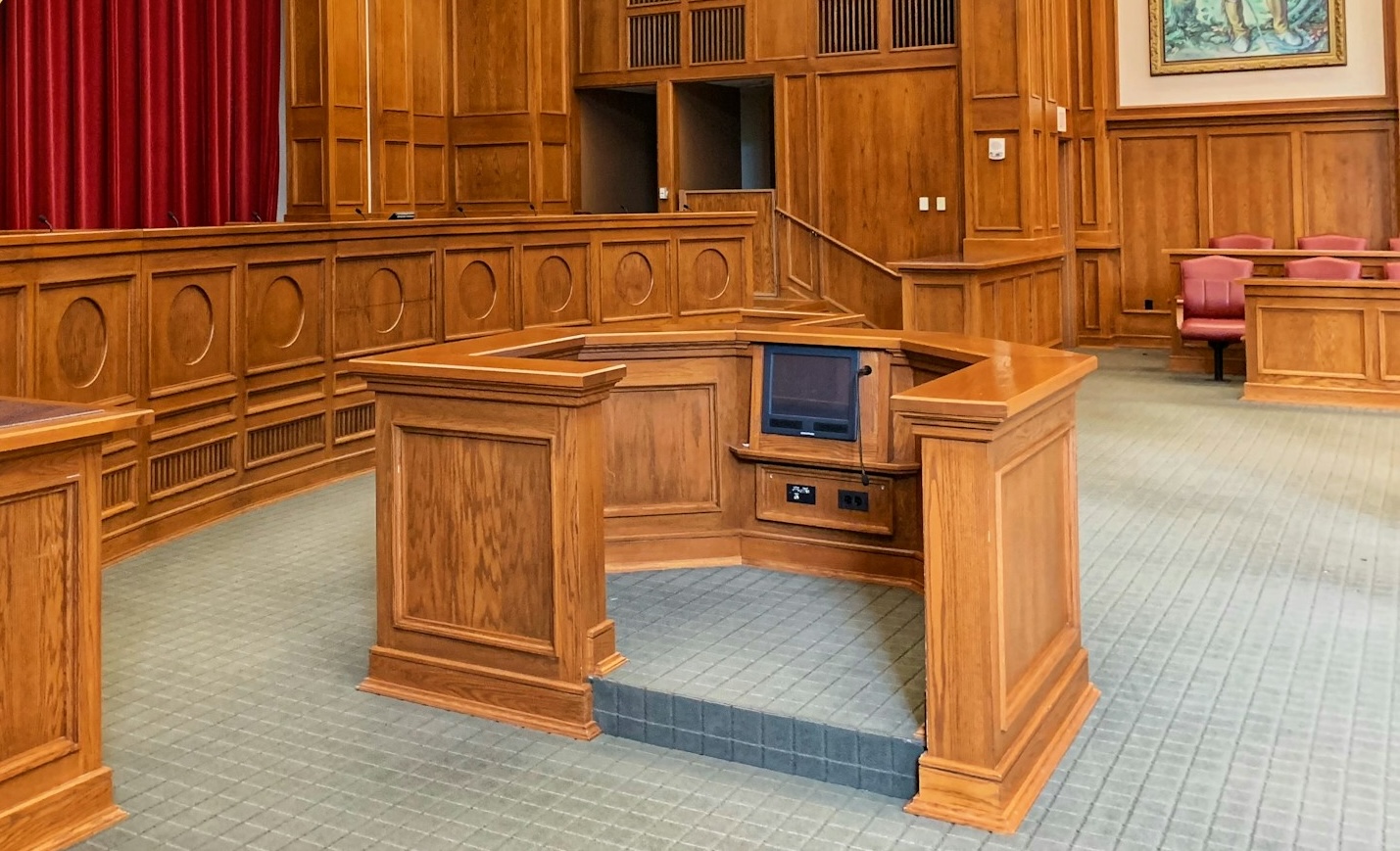 Interior of a courtroom witness stand with wooden paneling and a small monitor.