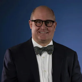 Portrait of a bald man wearing glasses, a suit, and a patterned bow tie against a dark blue background.