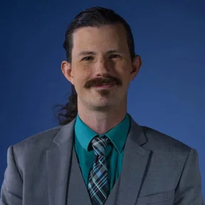 Portrait of a man with a mustache and long hair, wearing a gray suit and teal shirt.
