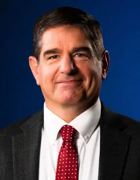 Portrait of a middle-aged man in a dark suit, white shirt, and red patterned tie against a blue background.