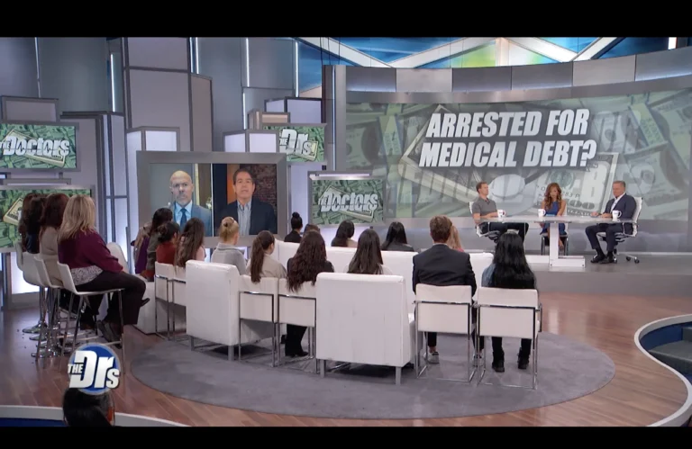 Television studio set of "The Doctors" with audience, hosts, and large screen reading "Arrested for Medical Debt?